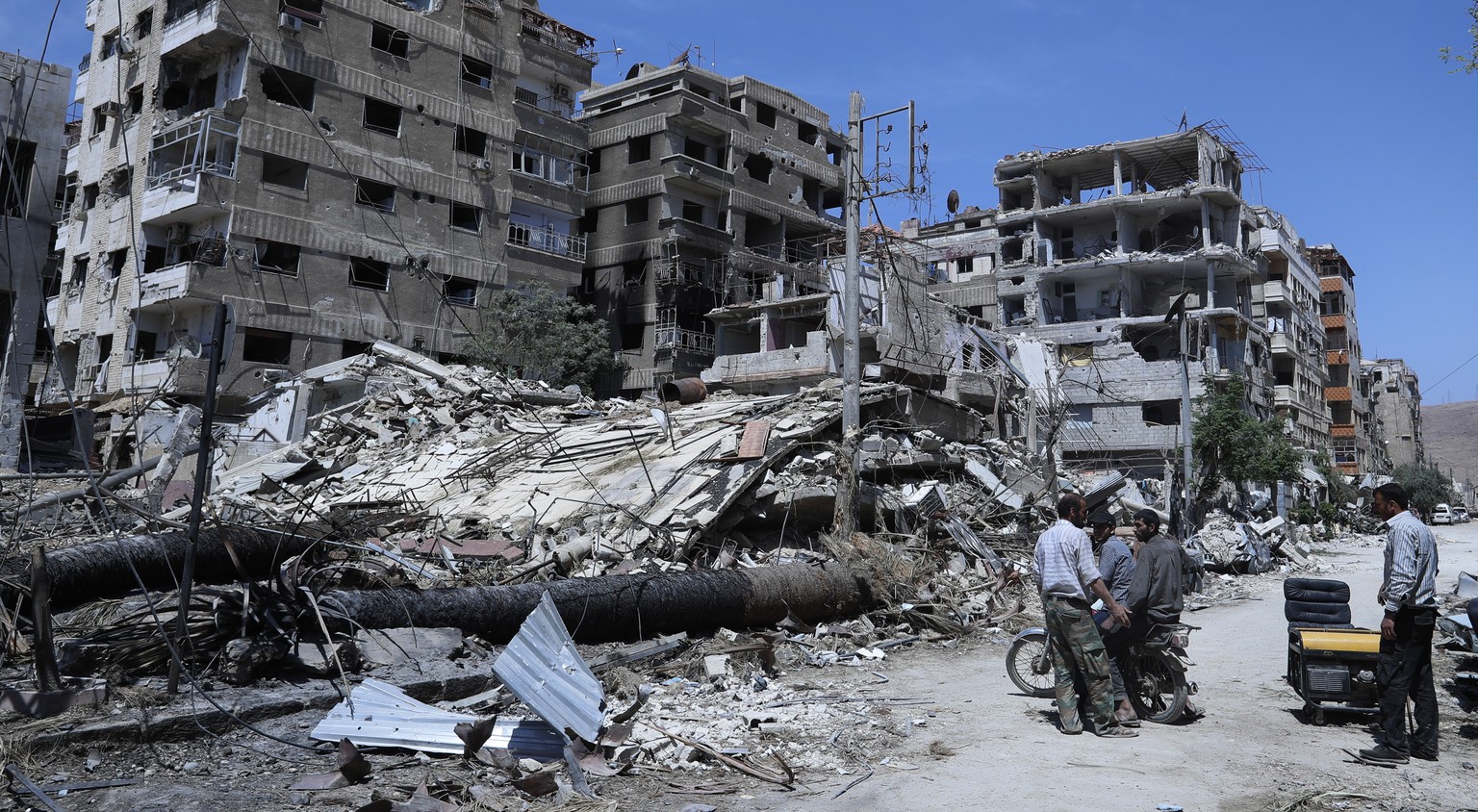 FILE - In this Monday, April 16, 2018 file photo, people stand in front of damaged buildings, in the town of Douma, the site of a suspected chemical weapons attack, near Damascus, Syria. The global ch ...