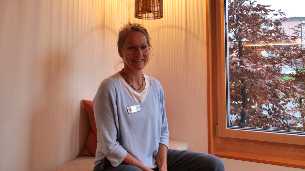 Christa Zumstein ist palliative Pflegefachfrau im Hospiz Zentralschweiz. Hier sitzt sie auf einem Sofa neben einem Fenster und lächelt in die Kamera.