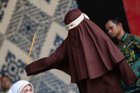 A shariah law official swings a rattan cane at a woman convicted of violating Islamic law by kissing, without being married, during a TikTok livestream, in Banda Aceh, Indonesia, Thursday, July 2, 202 ...