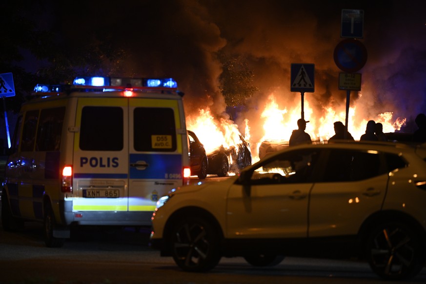epa10839218 People are silhouetted against the flames near police vehicles after a large number of cars were set on fire on Ramels vag in Rosengard, in Malmo, Sweden, 04 September 2023. According to t ...