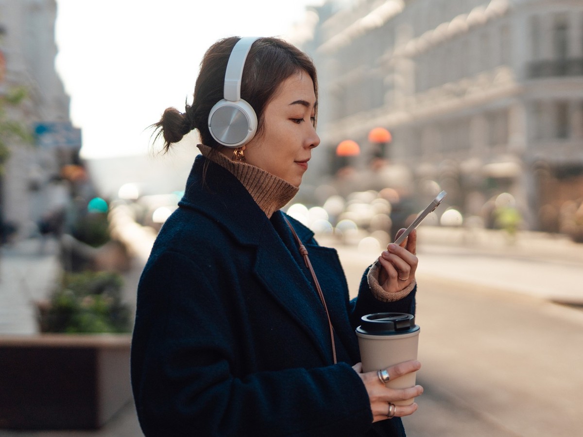 Young Asian woman looking at her mobile phone, wearing headphones, holding a takeaway coffee cup, standing in the city with warm sunlight, feeling relaxed and thoughtful during a commute. A quiet, per ...
