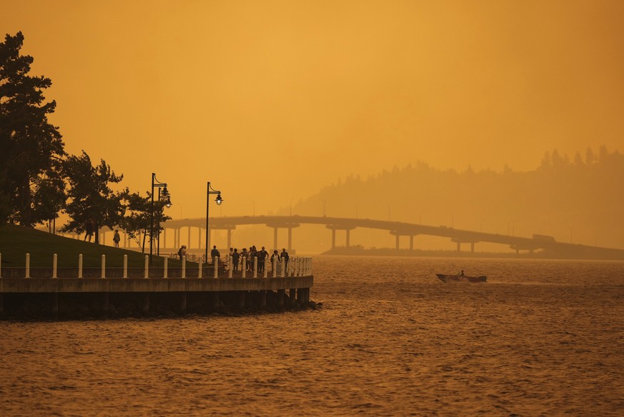 A person travels in a boat past people walking on the boardwalk as smoke from the McDougall Creek wildfire blankets the area on Okanagan Lake in Kelowna, British Columbia, Friday, Aug. 18, 2023. (Darr ...
