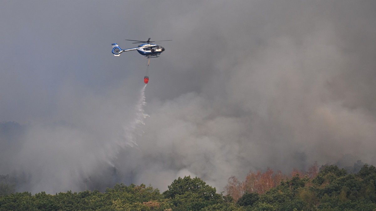 14.08.2026, Nordrhein-Westfalen, Hürtgenwald: Ein Polizeihubschrauber mit Löschwasser-Außenlastbehälter unterstützt die Löscharbeiten beim Waldbrand im Hürtgenwald. Mehrere Hundert Hektar Waldfläche s ...