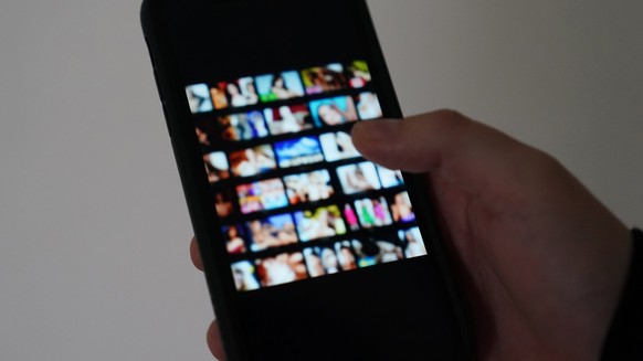 ILLUSTRATION - 19.10.2023, Hamburg: ARCHIV - Ein Mann schaut sich auf einem Smartphone eine Porno-Internetseite an (gestellte Szene). Es beginnt mit einem vermeintlichen Online-Flirt und endet mit Gel ...