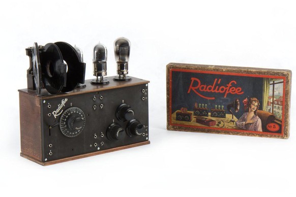 Röhrenradio «Radiofee II» zum Selbstbau, 1923. Die Abbildung auf der Schachtel gibt einen guten Eindruck vom Radiohören in den 1920er Jahren. Die Antenne hängt draussen, gehört wird via Kopfhörer oder Trichter-Lautsprecher. Elektrisch verstärkte Lautsprecher sind noch nicht auf dem Markt.