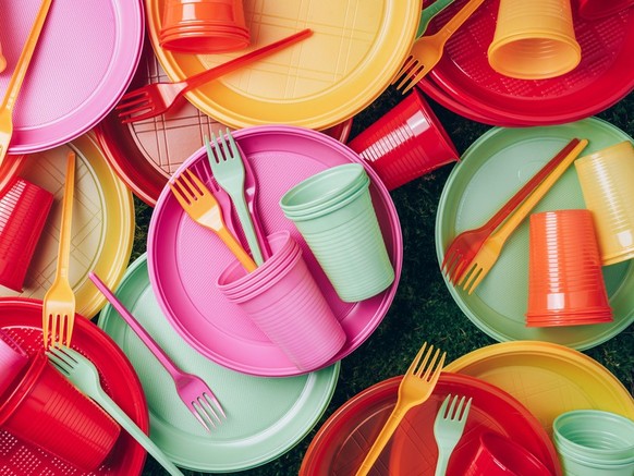 Disposable plastic tableware - plates, forks, spoons. Colored plastic disposable tableware on green grass moss background. Top view. Copy space. Birthday picnic utensil. Recycling plastic and ecology.