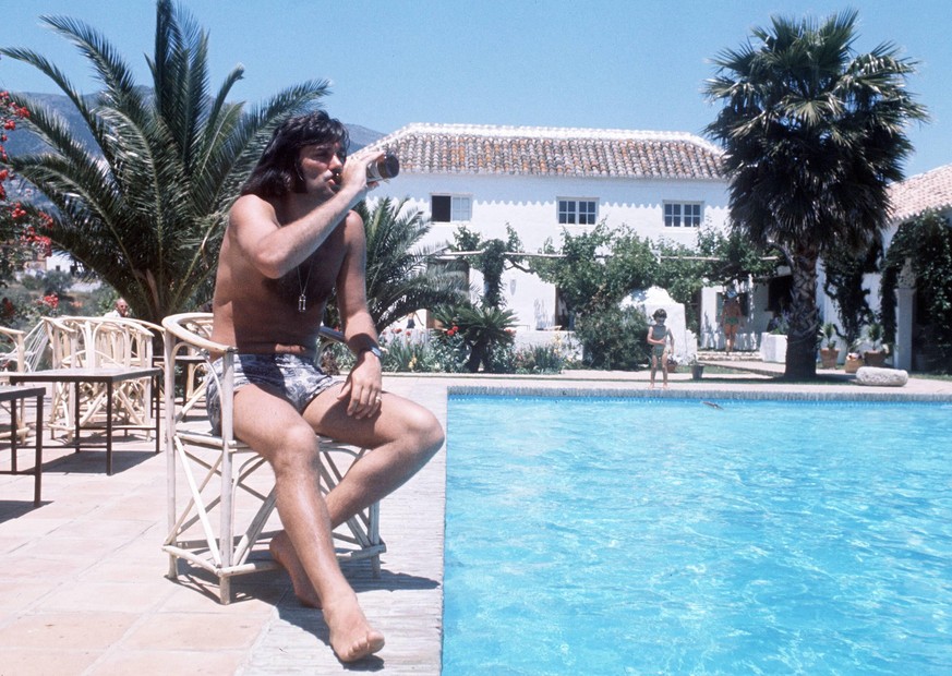 George Best (Nordirland) sitzt am Pool und nimmt einen Schluck aus einer Bierflasche

George Best Northern Ireland sits at Pool and Notes a Sip out a Beer bottle