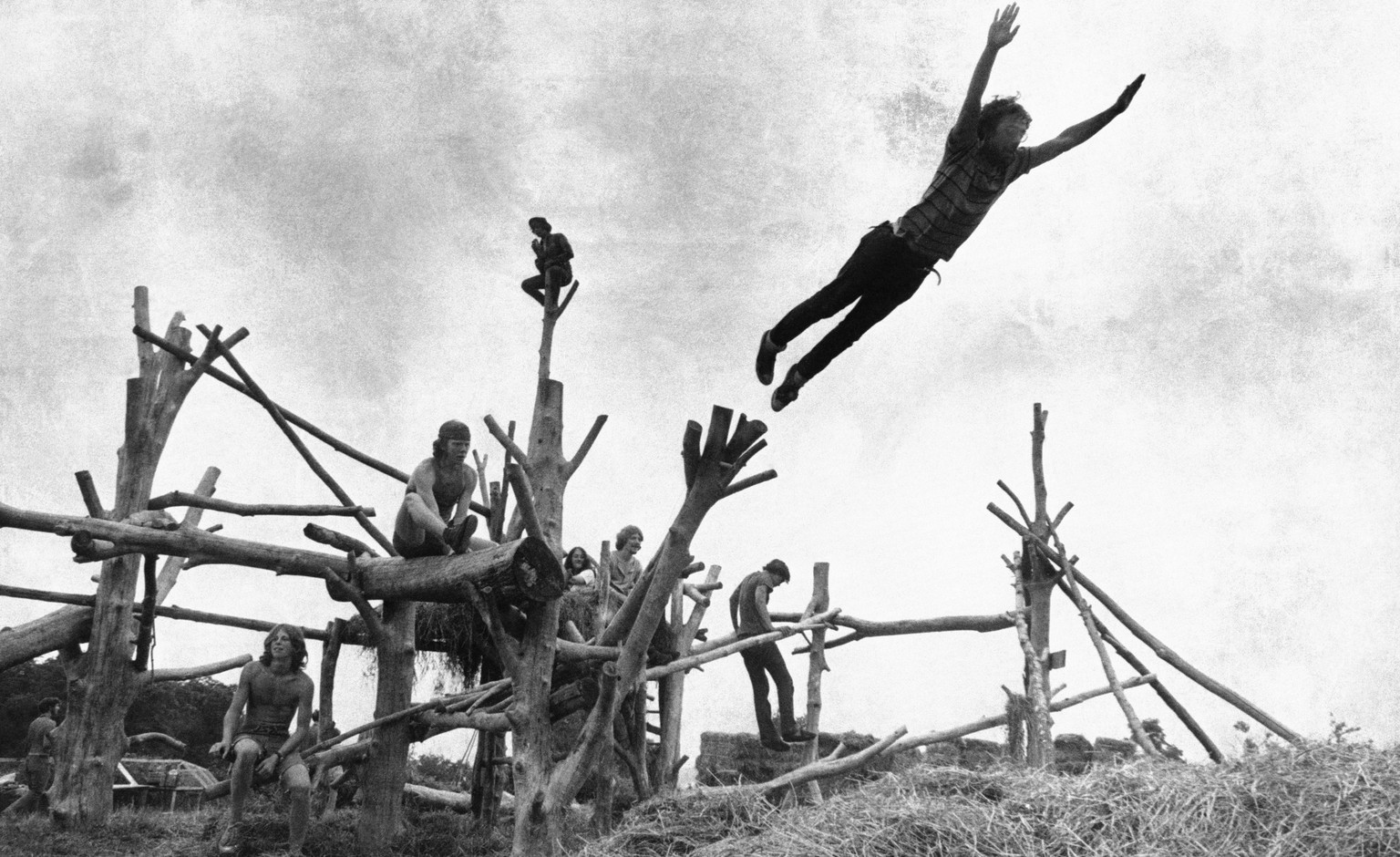 In this Aug. 15, 1969 file photo rock music fans sit on a tree sculpture as one leaps mid-air onto a pile of hay during the Woodstock Music and Art Festival held in Bethel, N.Y. To some Americans, the ...