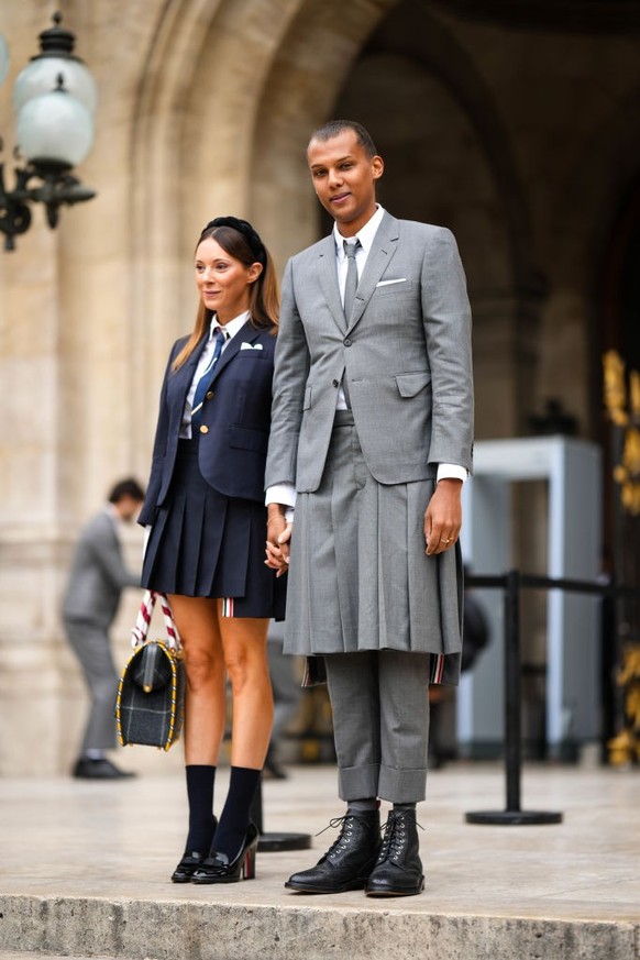 PARIS, FRANCE - OCTOBER 03: Stromae (L) wears a white shirt, a gray tie, a gray blazer jacket, a gray pleated / accordion knees skirt, matching gray suit pants, black shiny leather laces ankle boots,  ...