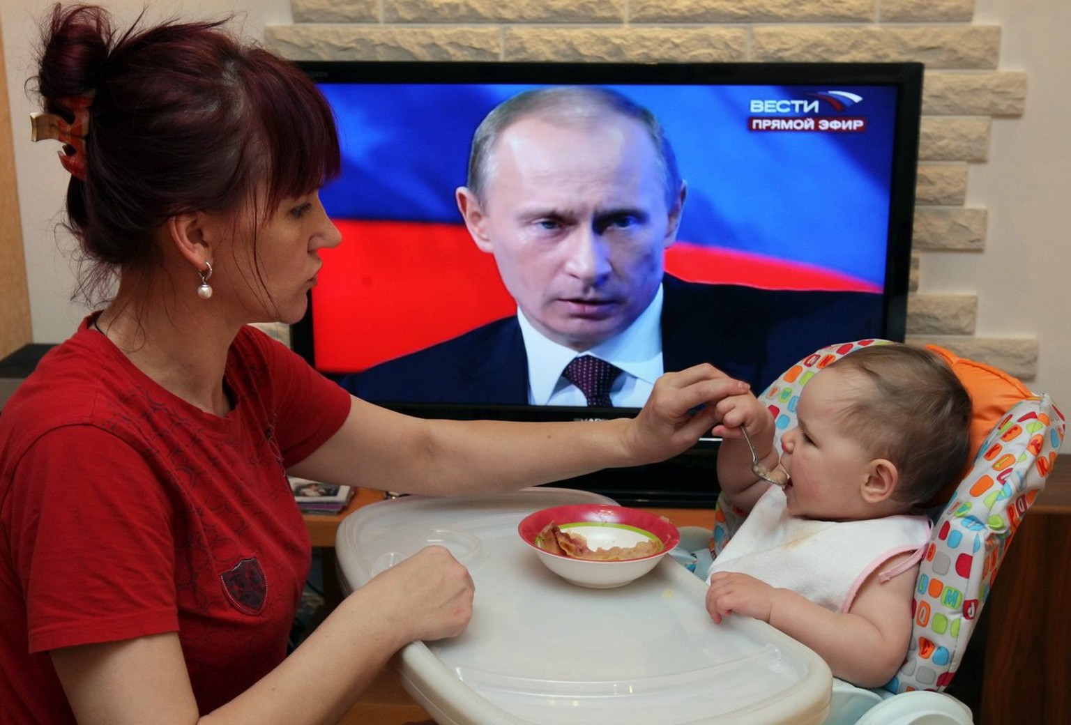 epa01953485 A woman spoon-feed a baby as the television displays Russian Premier Vladimir Putin during live call-in show, in Moscow, Russia, 03 December 2009. State news channels broadcasted the show. ...