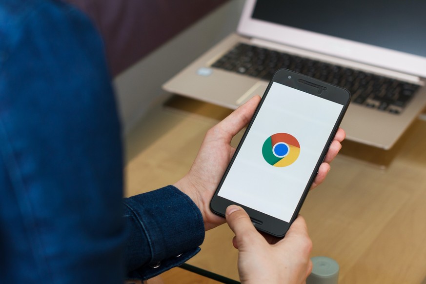 User mit Android-Smartphone und Google Chrome, im Hintergrund ein Laptop. Symbolbild