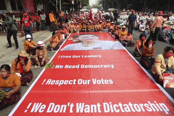 Protesters sit next to a giant banner with images of ousted leader Aung San Suu Kyi during an anti-coup protest in Mandalay, Myanmar, Sunday, Feb. 21, 2021. Police in Myanmar shot dead a few anti-coup ...