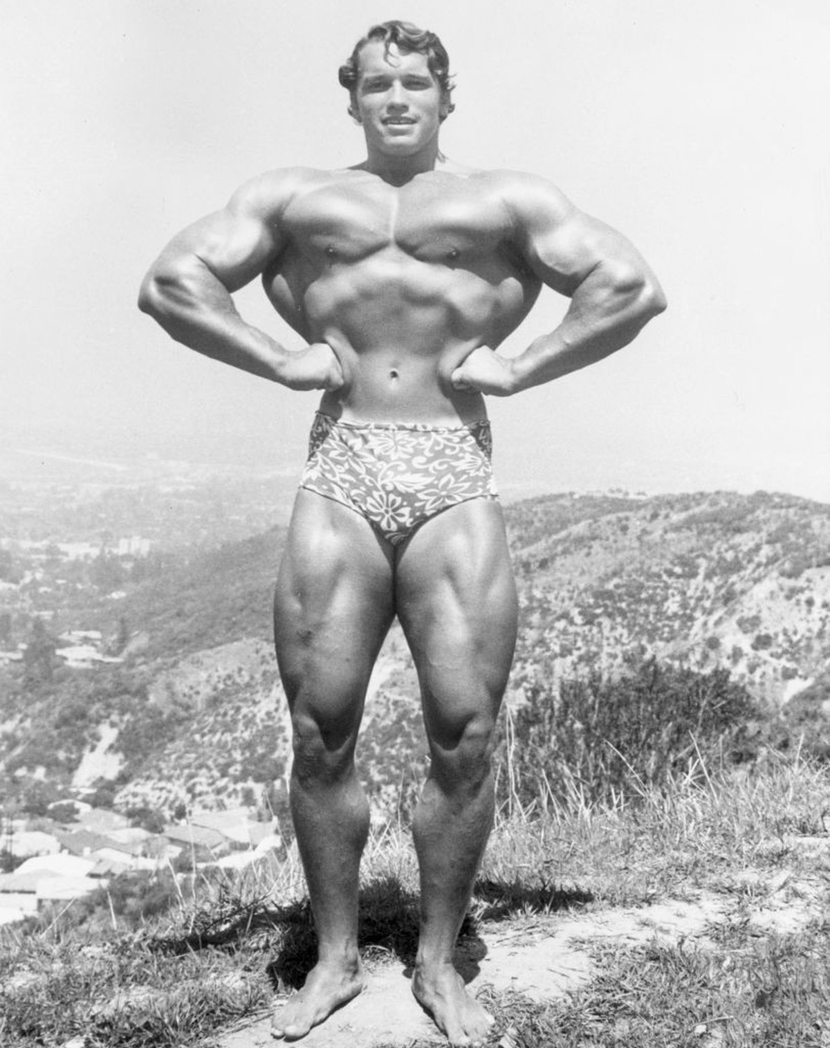 circa 1966: Full-length image of Austrian-born bodybuilder Arnold Schwarzenegger standing and flexing on top of a hillside near Muscle Beach, Santa Monica, California. He wears a floral print bathing  ...