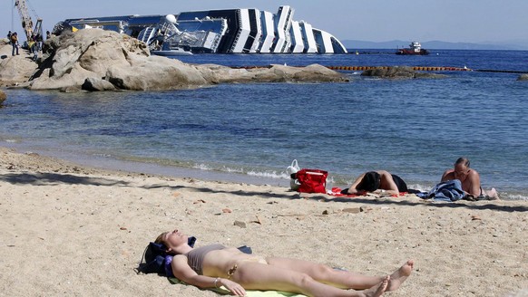 epa03176792 Tourists enjoy the weather and sun on the beach of Giglio Island on 09 April 2012 near the stricken cruise liner Costa Concordia on the background. The cruise liner rests half-sunken in fr ...