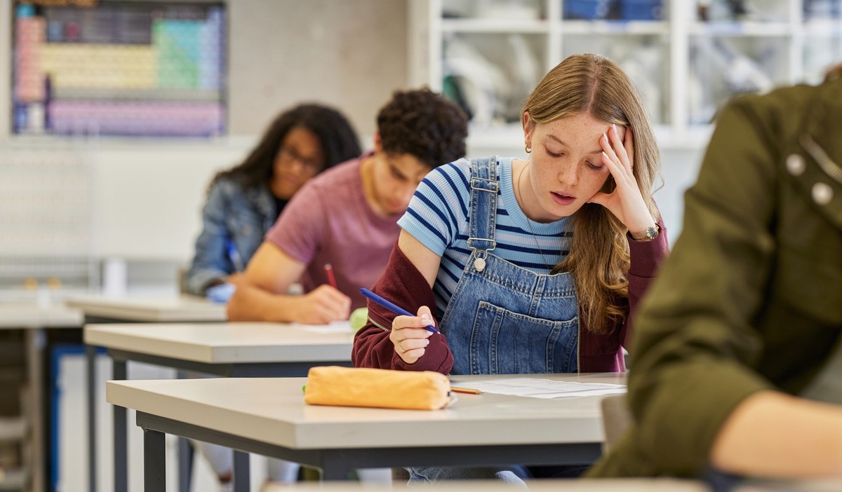 Stressed student holding head while taking exam in high school classroom. Tired girl showing signs of difficulty during test at secondary school with copy space. Classmates working silently while writ ...
