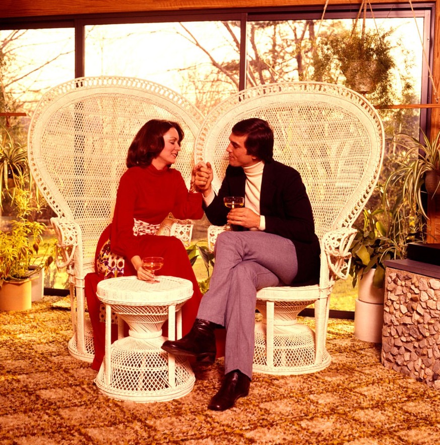29. Mai 1973 - MAN WOMAN COUPLE HOLDING HANDS DRINKING WINE ON PATIO SITTING WHITE WICKER RATTAN CHAIRS TROPICAL MOTIF (Photo by Photo Media/ClassicStock/Getty Images)