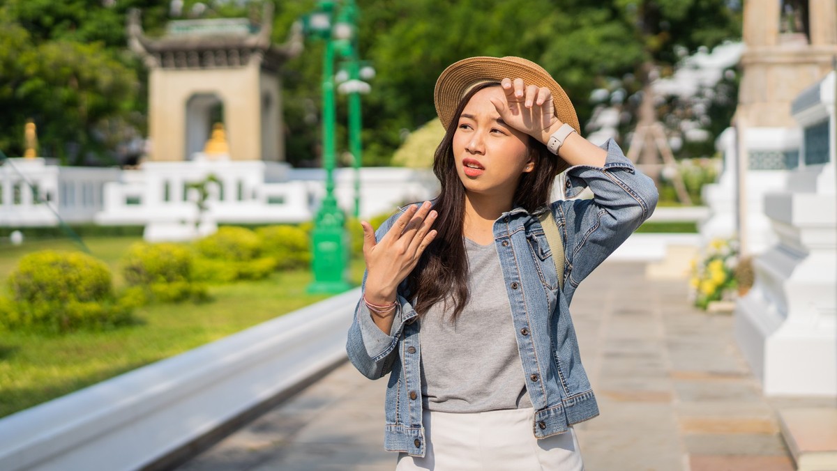 Portrait of Asian female traveler fanning and sweating on sidewalk of temple on street in Bangkok, Thailand, Southeast Asia in hot weather and temperature. Heatstroke Outdoor health problem concept xk ...