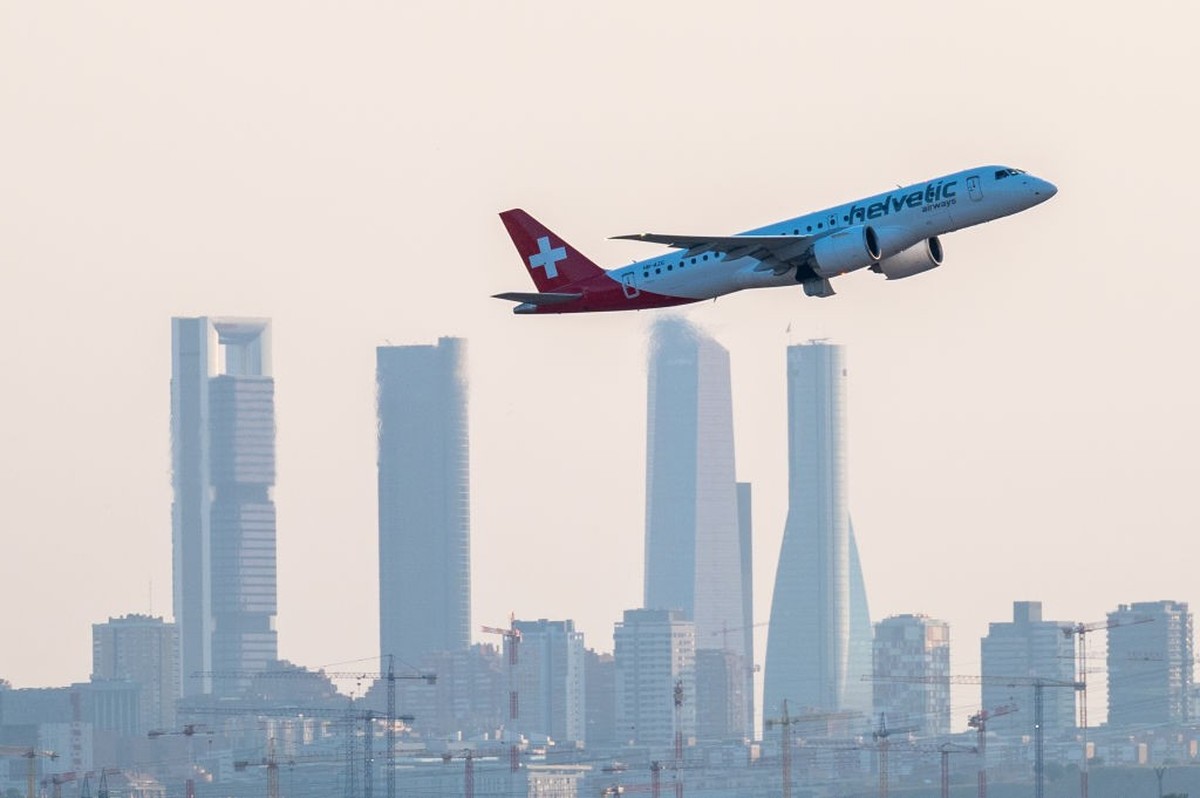 MADRID, SPAIN - 2023/07/22: A Swiss low cost Helvetic Airways comercial flight flies passing by the skyscrapers of Madrid's skyline, known as the "Four Towers Business Area", after take ...