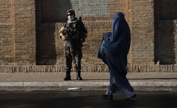 epa09354209 An armed Afghan security official stands guard as a woman wering a burqa walks by during Eid al-Adha prayers at a Mosque, in Herat, Afghanistan, 20 April 2021. The Afghan government and th ...