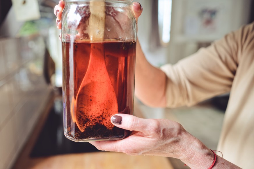 Both hands lift a large glass jar filled with deep amber sweetened tea toward the light, revealing loose tea leaves suspended inside, with a bright kitchen window providing a luminous backlit effect.  ...