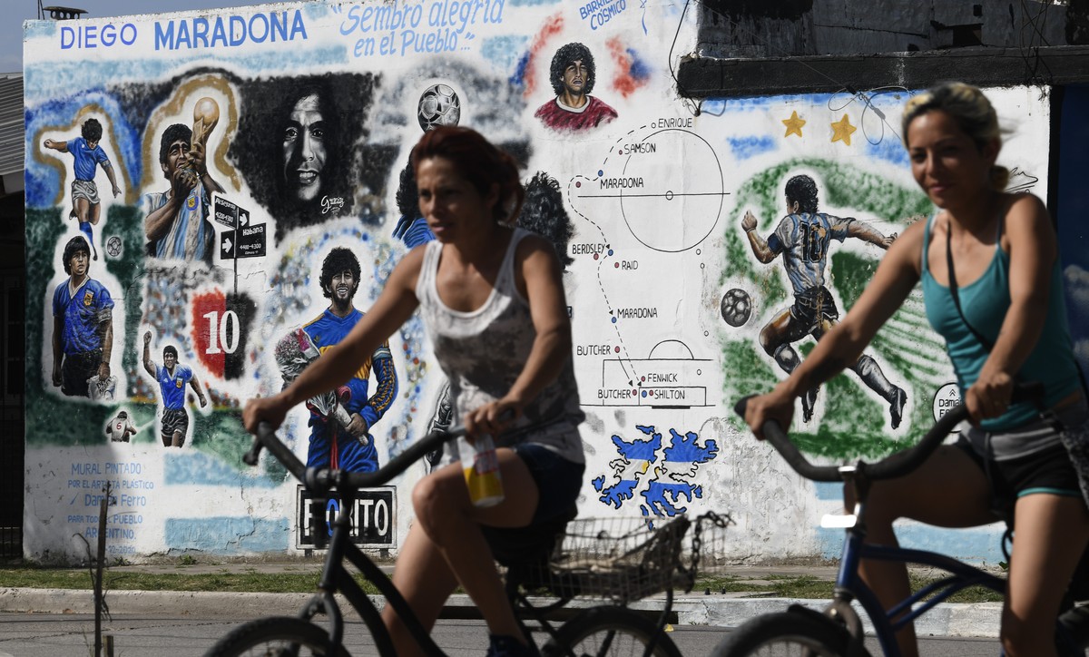 Women cycle past a mural depicting the late soccer legeng Diego Maradona in Lomas de Marilo neighborhood, Buenos Aires, Argentina, Monday, Nov. 22, 2021. Thursday marks the first anniversary of the so ...