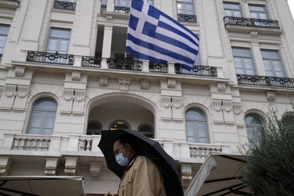 A man wearing a face mask to protect against coronavirus, walks outside an electronic store as a Greek flag waves in Athens, Greece, Tuesday, Nov. 2, 2021. Greek Health Minister will announce new measures against pandemic after Greece reached new record of daily cases with 5,449 on Monday. (AP Photo/Thanassis Stavrakis)