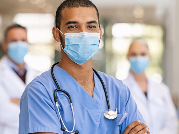 Confident multiethnic male nurse in front of his medical team looking at camera wearing face mask during covid-19 outbreak. Happy and proud indian young surgeon standing in front of his colleagues wea ...