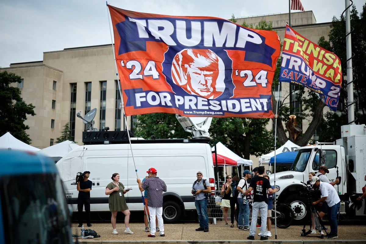 WASHINGTON, DC - AUGUST 03: A supporter carries a large flag in support of Donald Trump television satellite trucks outside the E. Barrett Prettyman U.S. District Court House ahead of Trump's arr ...