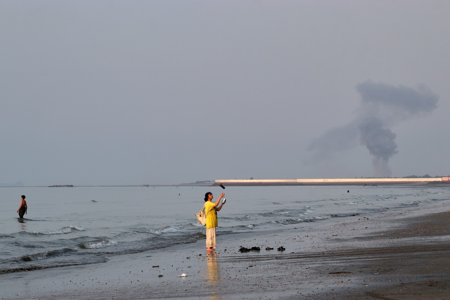 KEYPIX - A woman stands at the water's edge along the Strait of Hormuz as a plume of smoke rises in the background following an explosion, off Bandar Abbas, Iran, Monday, July 13, 2026. (KEYSTONE ...