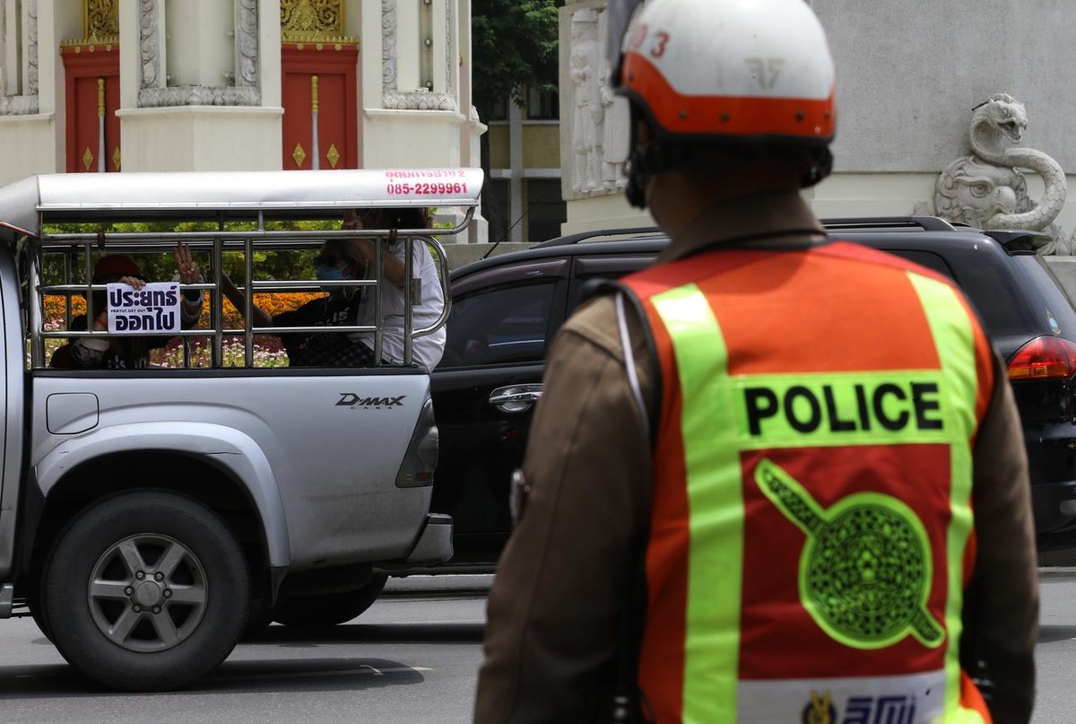 epa09335052 A Thai police officer keeps watch as anti-government protesters sitting in the back of a pickup truck display a placard reading 'Prayut get out' (L) during a car mob rally along  ...