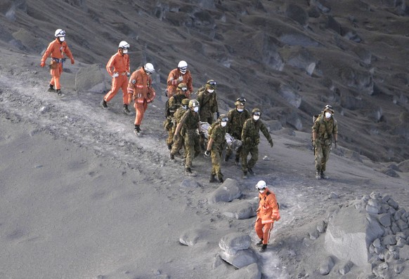 Rettungskräfte tragen verletzte Wanderer durch aschenbedeckte Wege den Vulkan herunter.