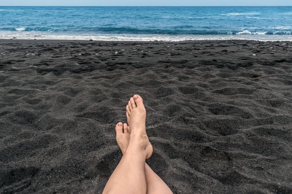 Woman feet lie on the black sand against the backdrop of the Atlantic Ocean in Tenerife, Spain. Relax on a black sand beach in the Canary Islands
