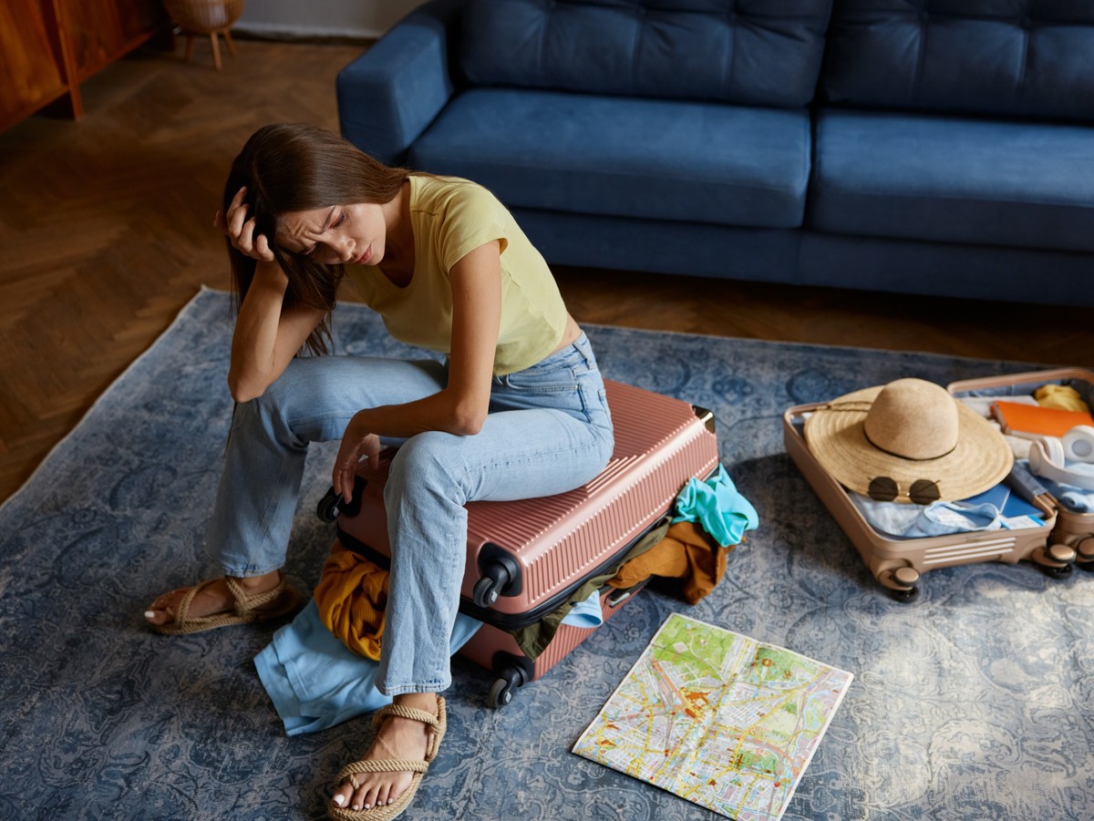Frustrated young woman sitting on travel suitcase with clothes excess, Frustrated young woman sitting on travel suitcase with clothes excess. Exhausted female traveler gesturing with irritation showin ...