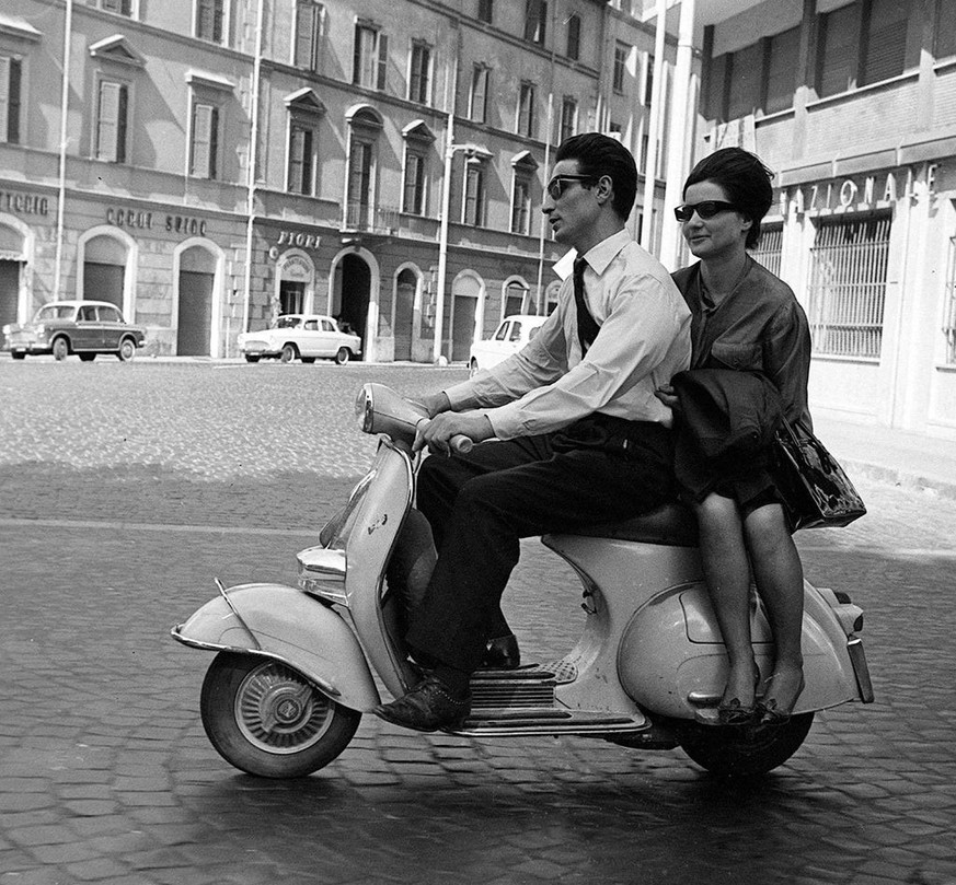 An everyday scene from post-war Italy. A pillion passenger rides side-saddle - a common sight before crash helmets became compulsory - while aftermarket side-panel footrests reflect the many practical ...