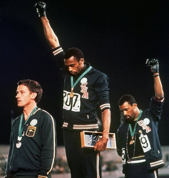 Extending gloved hands skyward in racial protest, U.S. athletes Tommie Smith, center, and John Carlos, stare downward during the playing of the Star Spangled Banner after Smith received the gold and C ...