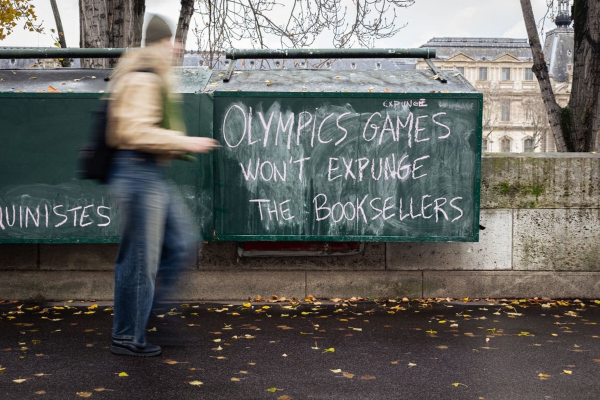 PARIS, FRANCE - 2023/12/13: A woman walks by a bookseller's stand with protest graffiti reading "Olympic Games won't expunge the booksellers". The famous booksellers (known as bouq ...
