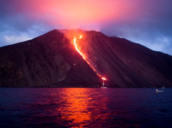 View of the Stromboli volcano from the sea at night, with lava flowing to the sea and its red reflected in clouds. Some ships in the distance.
