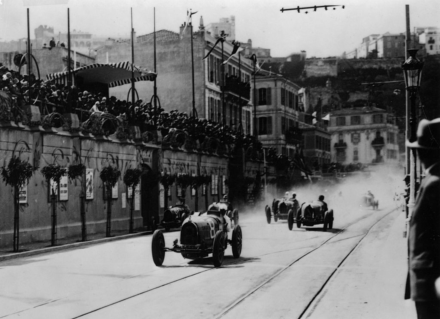 Start of the inaugural Monaco Grand Prix, 1929. Crowds watch as the cars speed along the street, led by a Bugatti. Note the tramlines. This first running of the event was won by &#039;W Williams&#039; ...