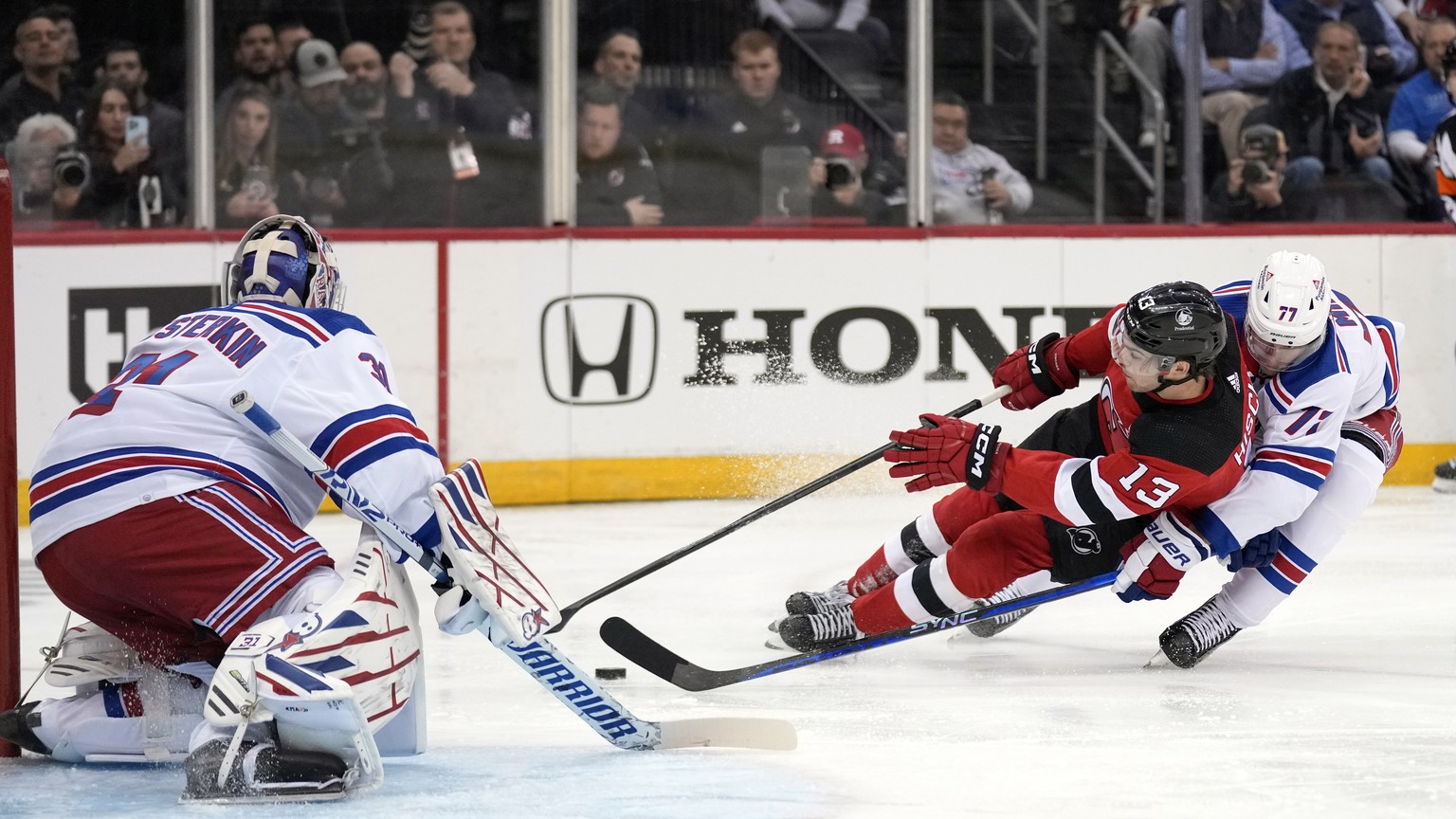 New Jersey Devils&#039; Nico Hischier (13), center, slides as he tries to make a shot on New York Rangers&#039; goaltender Igor Shesterkin, left, while being defended by Niko Mikkola, right, during th ...