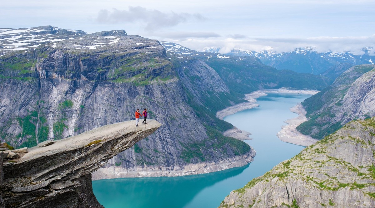 Trolltunga, Norway, A View From The Edge, couple of men and women on top of a mountain edge Two people stand on the edge of Trolltunga, Norway, a cliff overlooking a serene Norwegian lake and the surr ...