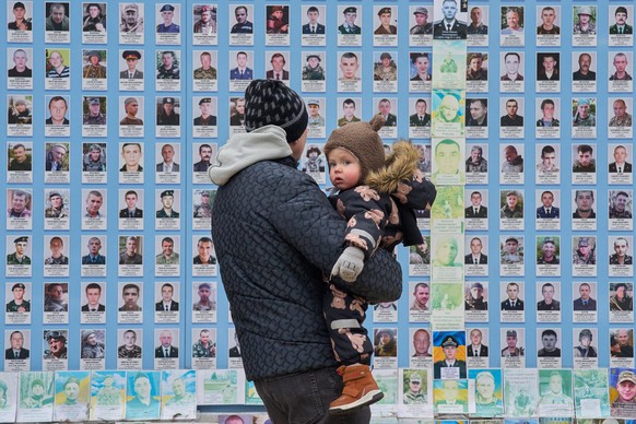 A man holds his child at the Memorial Wall of Fallen Defenders of Ukraine in Russian-Ukrainian War and shows photos of his killed fellow soldiers in Kyiv, Ukraine, Thursday, Dec. 25, 2025. (AP Photo/E ...