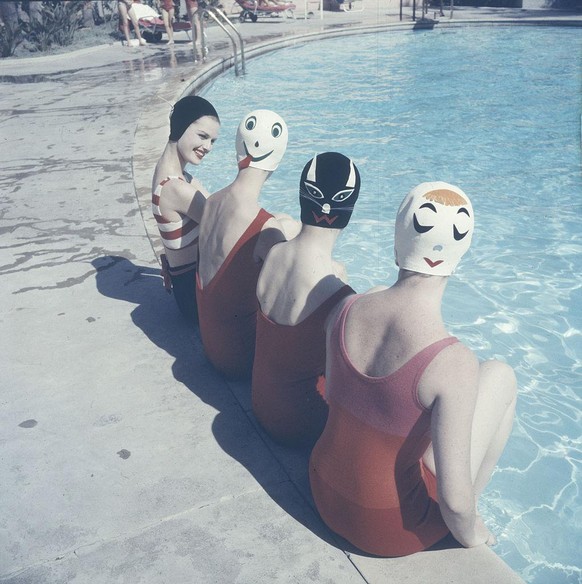 Four women sits on the deck of a swimming pool, their legs in the water, and model bathing caps that feature various fanciful faces, March 1959. Invented by Long Island, New York, resident Betty Geib, ...